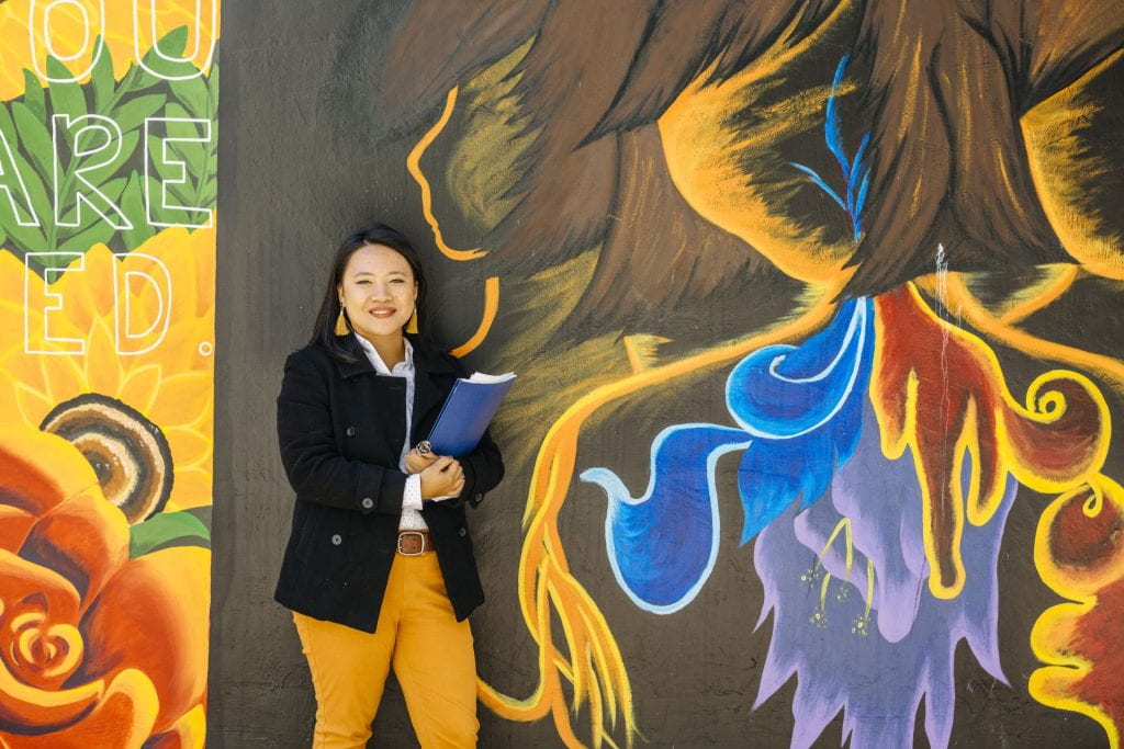 Smiling female student is posed with notebook in hand, in front of a colorful mural.  